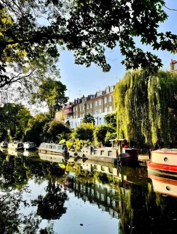 A tranquil canal scene with moored narrowboats. The canal is lined with lush greenery, including large trees and a weeping willow, with a row of traditional brick houses in the background.