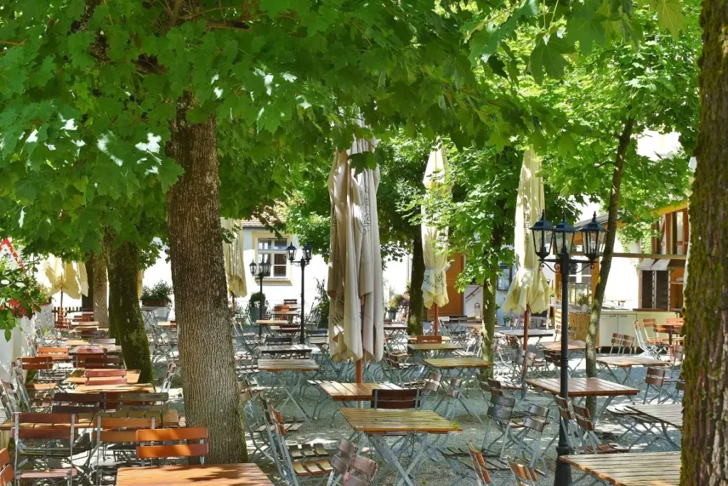 An outdoor beer garden featuring numerous tables and chairs arranged under the shade of large green trees. The area is equipped with folded beige umbrellas and decorative black lamp posts.