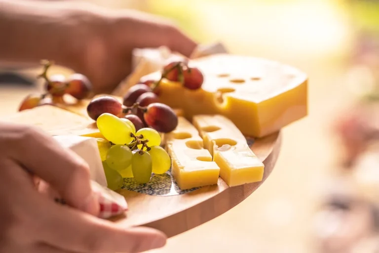Close-up of a cheese platter with various types of cheese, including Swiss cheese with holes, accompanied by red and green grapes. The platter is being held by a person, with a soft, warm background light.