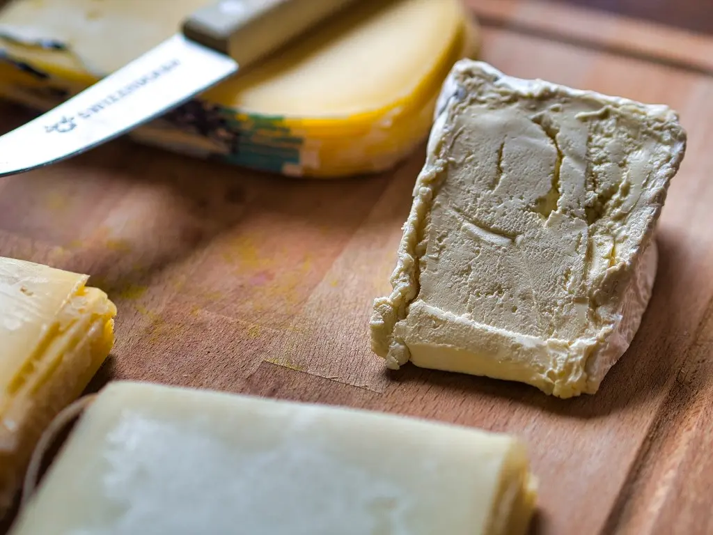 Close-up of various cheeses on a wooden cutting board. The selection includes a soft, rind-covered cheese and slices of a firmer yellow cheese. A cheese knife is visible in the background.