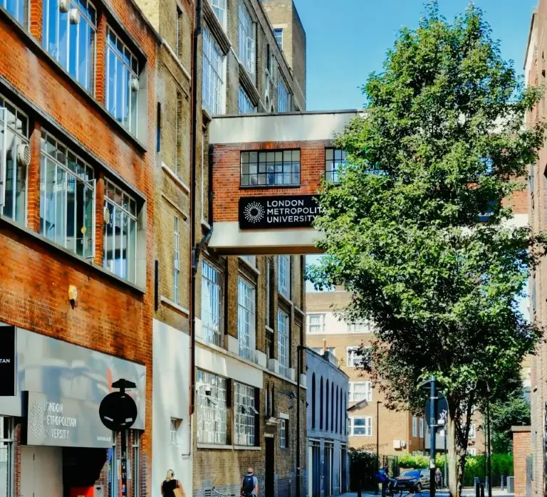 Street view of London Metropolitan University with a bridge connecting two buildings, featuring a sign with the university's name. The scene includes pedestrians, bicycles parked along the sidewalk, and a tree providing shade.