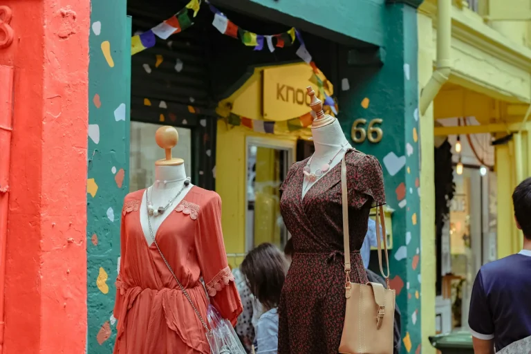 Mannequins displaying vintage dresses outside a colorful boutique, featuring a coral dress and a brown patterned dress with accessories. The store's entrance has vibrant painted walls and decorative bunting above the door.