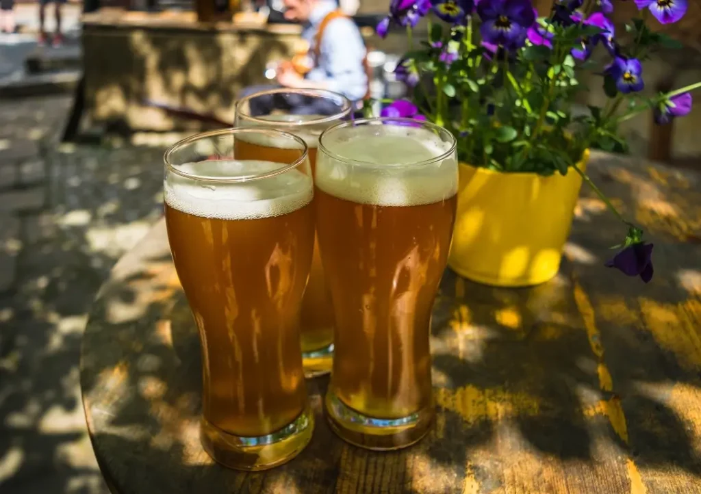 hree glasses of wheat beer with a foamy head, arranged on a wooden outdoor table in a sunny setting. A pot of purple flowers in a yellow planter adds a touch of color to the scene, with blurred figures and shadows in the background.
