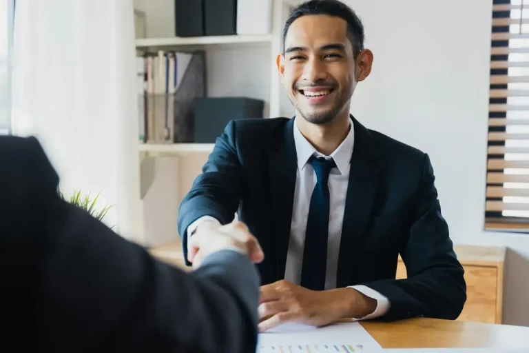 A smiling man in a suit is shaking hands with another individual in a professional setting after agreeing a positive outcome.
