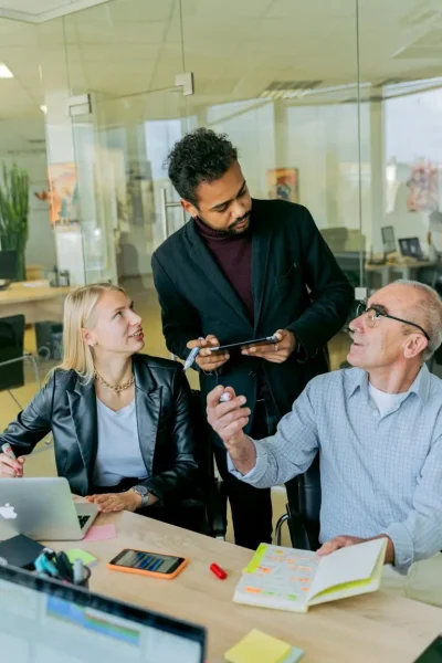 Three colleagues are gathered in a modern office setting, exchanging ideas. One person is holding a tablet, while the others are attentively listening.